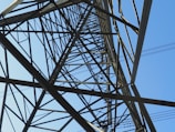 An intricate network of metal beams forms the structure of a transmission tower, viewed from below. The geometric lines and angles of the steel lattice create a complex pattern against a clear blue sky.