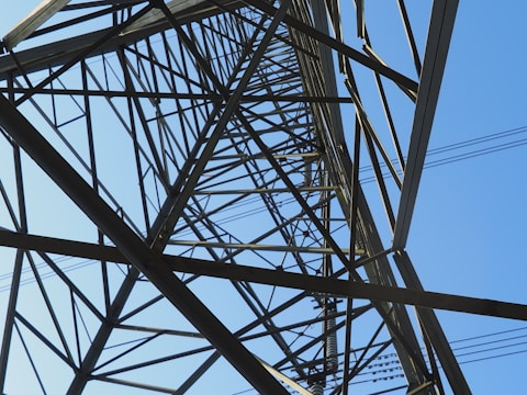 An intricate network of metal beams forms the structure of a transmission tower, viewed from below. The geometric lines and angles of the steel lattice create a complex pattern against a clear blue sky.