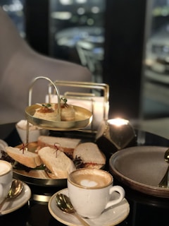 A two-tiered serving platter featuring assorted sandwiches and pastries. There are two cups of cappuccino, each on a saucer with a spoon, all set on a sleek, dark table. The background is softly blurred, suggesting a cozy, modern setting.