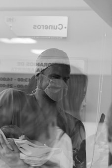 A person wearing a surgical mask and scrub cap is holding a newborn baby wrapped in a blanket. The scene appears to be through a glass or transparent partition with reflected images, creating a layered effect. The environment suggests a hospital or medical facility, evidenced by signage visible in the background.