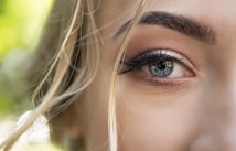 A close-up of a person's eye, showcasing detailed features such as eyelashes, eyebrows, and the eye's iris. The background is softly blurred, adding depth and emphasizing the eye. Light strands of hair are visible, adding an element of softness to the image.