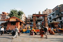 A bustling street scene features a traditional market with vibrant stalls displaying flowers and other items. The foreground includes people walking by and scooters parked nearby, while a prominent, intricately designed brick gate stands as the focal point. Behind the gate, a variety of colorful buildings and structures are visible, creating a lively urban atmosphere.
