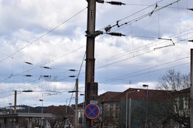 Overhead power lines are attached to a wooden pole in an urban area. Buildings with red-tiled roofs and a no parking sign are visible in the background, under a cloudy sky.