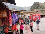 A vibrant marketplace scene with colorful textiles hanging from stalls on the left, featuring patterns in red, orange, and purple. Several people are interacting near a display of jewelry, while a large red Coca-Cola truck is parked in the background. Traditional architecture and mountainous scenery form the backdrop.