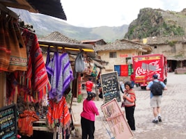 A vibrant marketplace scene with colorful textiles hanging from stalls on the left, featuring patterns in red, orange, and purple. Several people are interacting near a display of jewelry, while a large red Coca-Cola truck is parked in the background. Traditional architecture and mountainous scenery form the backdrop.