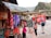 A vibrant marketplace scene with colorful textiles hanging from stalls on the left, featuring patterns in red, orange, and purple. Several people are interacting near a display of jewelry, while a large red Coca-Cola truck is parked in the background. Traditional architecture and mountainous scenery form the backdrop.