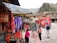 A vibrant marketplace scene with colorful textiles hanging from stalls on the left, featuring patterns in red, orange, and purple. Several people are interacting near a display of jewelry, while a large red Coca-Cola truck is parked in the background. Traditional architecture and mountainous scenery form the backdrop.