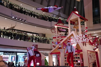A festive indoor scene depicting a holiday performance. A performer, dressed in a Santa-themed outfit, is airborne with dynamic acrobatic form above a stage decorated as 'Santa Academy.' The stage, designed like a wooden house with gabled roofs and a bell tower, is adorned with red and white holiday-themed elements. Additional performers in similar attire are performing on the stage below. An audience gathers on multiple levels of the mall, lined with garland and lights, observing the performance.