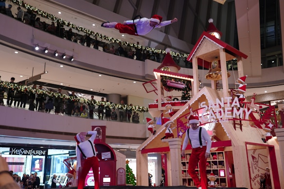 A festive indoor scene depicting a holiday performance. A performer, dressed in a Santa-themed outfit, is airborne with dynamic acrobatic form above a stage decorated as 'Santa Academy.' The stage, designed like a wooden house with gabled roofs and a bell tower, is adorned with red and white holiday-themed elements. Additional performers in similar attire are performing on the stage below. An audience gathers on multiple levels of the mall, lined with garland and lights, observing the performance.
