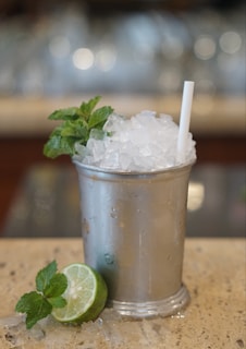 A refreshing drink served in a metallic cup, filled with crushed ice and garnished with fresh mint leaves. A straw is inserted into the ice, and a lime half with more mint sits on the granite surface nearby. The background is softly blurred, emphasizing the beverage.