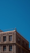 An urban building with a classic architectural design, featuring rows of windows and detailed brickwork. A solitary security camera is mounted on the corner of the building's rooftop, with a clear blue sky as the backdrop.