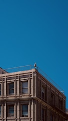 An urban building with a classic architectural design, featuring rows of windows and detailed brickwork. A solitary security camera is mounted on the corner of the building's rooftop, with a clear blue sky as the backdrop.