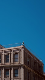 An urban building with a classic architectural design, featuring rows of windows and detailed brickwork. A solitary security camera is mounted on the corner of the building's rooftop, with a clear blue sky as the backdrop.
