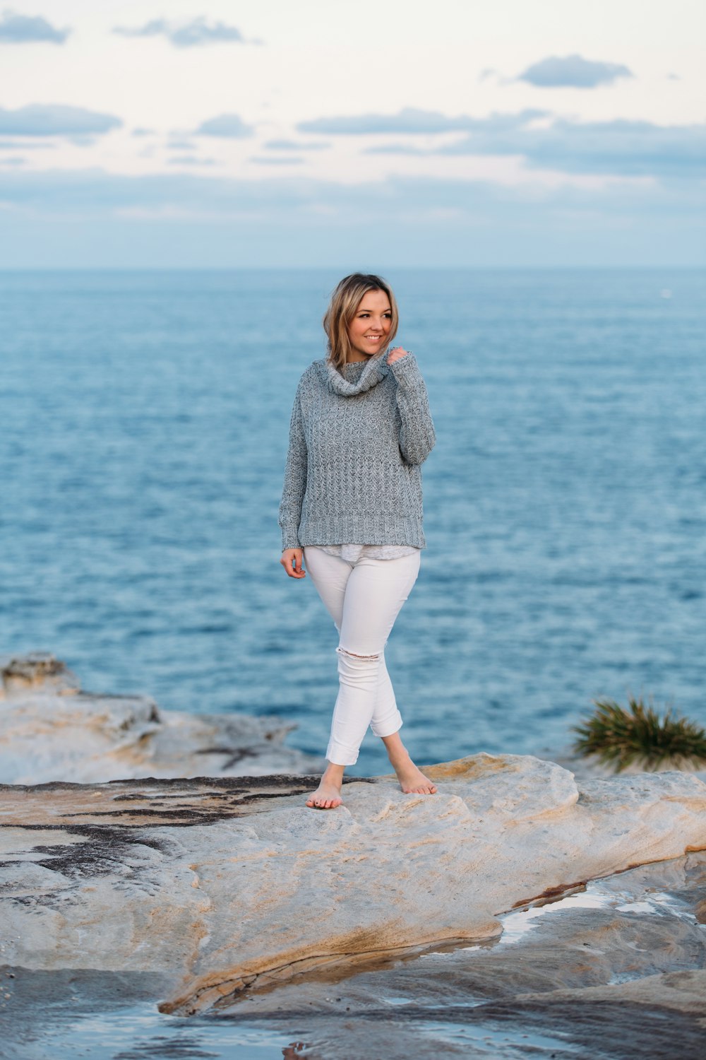 woman in gray sweater and white pants standing on brown rock near body of water during