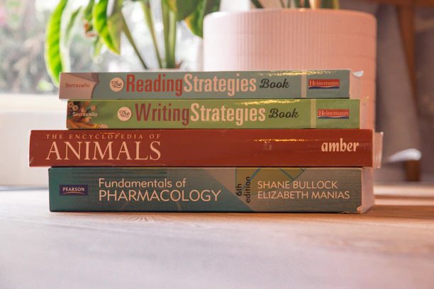 Stack of elegant books with serif fonts on a cream-colored wooden table near a green plant.