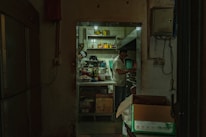 A dimly lit, compact kitchen is visible through an open doorway. Two people are inside, one busy at a counter with various cooking ingredients and utensils scattered around. Shelves hold condiments and kitchen supplies. The walls are slightly worn, adding a rustic feel to the environment.