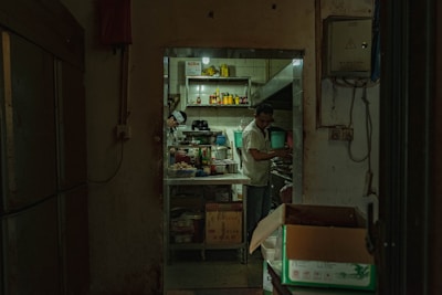 A dimly lit, compact kitchen is visible through an open doorway. Two people are inside, one busy at a counter with various cooking ingredients and utensils scattered around. Shelves hold condiments and kitchen supplies. The walls are slightly worn, adding a rustic feel to the environment.