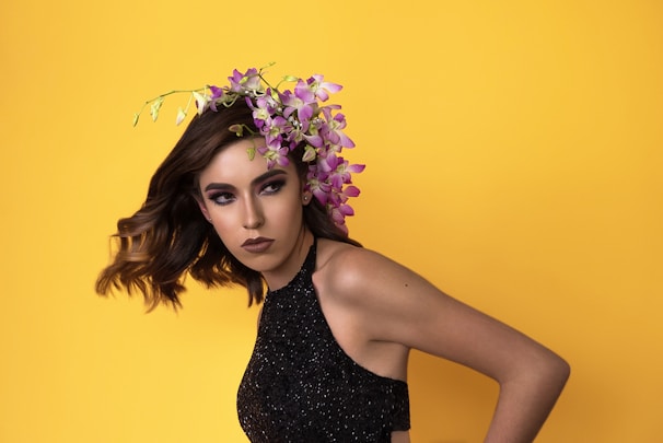 A person with wavy brown hair is wearing a black sequined dress. An arrangement of purple and pink flowers is placed in their hair, and they are set against a bright yellow backdrop. Their makeup is bold, featuring dark eyeshadow and lipstick.