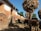 A rustic village setting features traditional mud-brick houses with thatched roofs, and livestock such as cows grazing in the foreground. Large bundles of corn are stacked on wooden structures, and a person is tending to the animals under a clear blue sky.