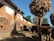A rustic village setting features traditional mud-brick houses with thatched roofs, and livestock such as cows grazing in the foreground. Large bundles of corn are stacked on wooden structures, and a person is tending to the animals under a clear blue sky.