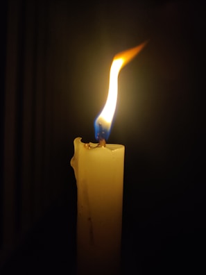 A single burning candle with a bright flame illuminates the dark surroundings. The wax on the candle appears slightly melted, showcasing a natural texture.