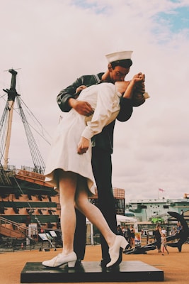 A large sculpture depicts a sailor kissing a nurse in an embrace reminiscent of the famous V-J Day in Times Square photograph. The figures are dressed in period clothing, with the sailor in navy attire and the nurse in a white uniform. The background features a historic ship and visitors walking around, suggesting an open-air museum or historical site.