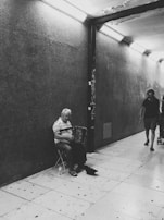 A black and white photograph of an older man sitting on a folding chair playing an accordion in a dimly lit, tiled underground passage. He is wearing a striped shirt and appears to be focused on playing his instrument. On the right side of the image, two other people are walking through the passage, one of them pushing a stroller.