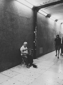 A black and white photograph of an older man sitting on a folding chair playing an accordion in a dimly lit, tiled underground passage. He is wearing a striped shirt and appears to be focused on playing his instrument. On the right side of the image, two other people are walking through the passage, one of them pushing a stroller.
