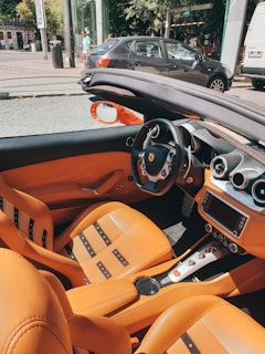 The interior of a luxury convertible car with tan leather seats, a polished dashboard, and a steering wheel featuring a prominent brand logo. The top is retracted, and the car is parked on a street with other vehicles and a few pedestrians in the background.