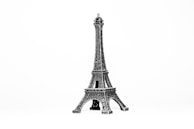 A small metallic replica of the Eiffel Tower is displayed against a plain white background. The intricate lattice work on the tower is clearly visible, showcasing detailed craftsmanship.