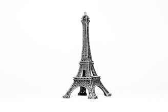 A small metallic replica of the Eiffel Tower is displayed against a plain white background. The intricate lattice work on the tower is clearly visible, showcasing detailed craftsmanship.