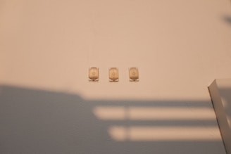 Three small, beige wall mounts arranged in a horizontal line on a solid light-colored wall with the shadow of a railing cast across the surface, creating a pattern of horizontal stripes.