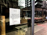 A storefront window with a sign from North Star Coffee Shop announcing closure for the foreseeable future. The sign thanks supporters and advises checking Instagram for reopening dates. Reflecting in the glass are outdoor tables and chairs, along with surrounding buildings and trees.