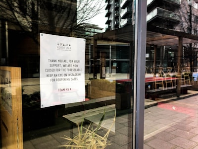A storefront window with a sign from North Star Coffee Shop announcing closure for the foreseeable future. The sign thanks supporters and advises checking Instagram for reopening dates. Reflecting in the glass are outdoor tables and chairs, along with surrounding buildings and trees.