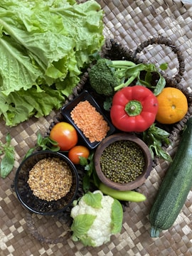 A vibrant assortment of fresh vegetables and grains is displayed on a woven mat. There are leafy greens, including lettuce and broccoli, and red lentils in a square bowl. A round red pepper and orange sit next to tomatoes, with a zucchini extending to the right. Small bowls contain grains and beans, and there is a head of cauliflower with light green leaves visible.