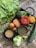 A vibrant assortment of fresh vegetables and grains is displayed on a woven mat. There are leafy greens, including lettuce and broccoli, and red lentils in a square bowl. A round red pepper and orange sit next to tomatoes, with a zucchini extending to the right. Small bowls contain grains and beans, and there is a head of cauliflower with light green leaves visible.