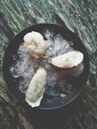 Three oysters topped with foam and a small garnish are placed on a bed of crushed ice within a dark bowl. The surface beneath has a marbled appearance with shades of green and white.
