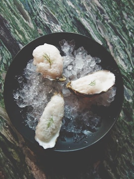 Three oysters topped with foam and a small garnish are placed on a bed of crushed ice within a dark bowl. The surface beneath has a marbled appearance with shades of green and white.