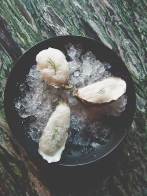 Three oysters topped with foam and a small garnish are placed on a bed of crushed ice within a dark bowl. The surface beneath has a marbled appearance with shades of green and white.