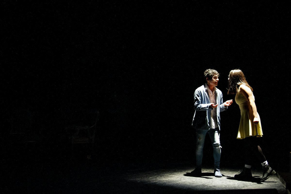 Two actors facing each other in a single white spotlight on a dark stage