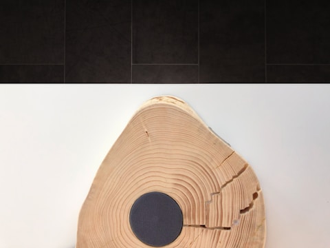 A piece of wood displaying prominent rings and natural cracks, with a circular, dark speaker or device embedded in its center. The wood rests on a smooth white surface against a dark, tiled background.