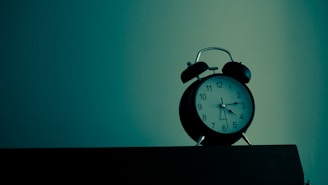 An analog alarm clock sits on a dark wooden surface with a teal and greenish background. The clock has a black body with a white clock face and black numbers. The lighting creates a moody and slightly mysterious atmosphere.