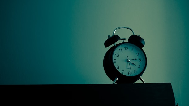 An analog alarm clock sits on a dark wooden surface with a teal and greenish background. The clock has a black body with a white clock face and black numbers. The lighting creates a moody and slightly mysterious atmosphere.
