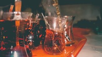 A series of curved glass cups filled with a dark beverage are placed on a tray. The beverage is poured into one of the glasses, creating a rich, bubbling effect. The setting appears dimly lit, focusing on the amber hues of the liquid and reflections on the glass surfaces.