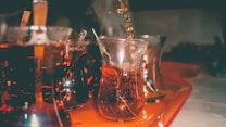 A series of curved glass cups filled with a dark beverage are placed on a tray. The beverage is poured into one of the glasses, creating a rich, bubbling effect. The setting appears dimly lit, focusing on the amber hues of the liquid and reflections on the glass surfaces.