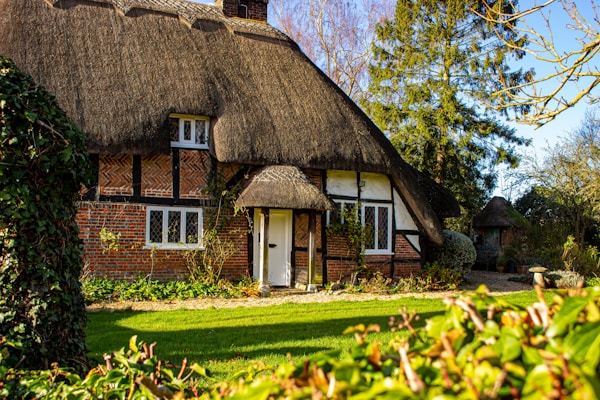 A quaint, traditional English cottage with a thatched roof is nestled in a lush garden. The brick and timber construction displays a charming, rustic appearance. A neat lawn and various shrubs surround the cottage, while tall trees provide a natural backdrop. The bright and clear sky adds to the serene setting.