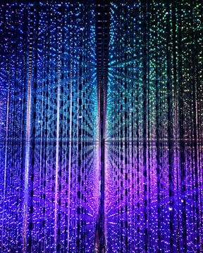 An array of vertical strings of lights creates a captivating, immersive display. The lights transition smoothly from purple at the bottom to blue and green at the top, forming a mesmerizing gradient effect. The arrangement gives a sense of depth and movement, as if exploring a futuristic, kaleidoscopic space.
