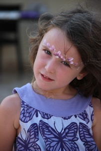 A young girl with long brown hair and a gentle expression. She has floral face paint in pink and white adorning her forehead and wears a sleeveless dress featuring a butterfly pattern in purple and white. The blurred background suggests an outdoor setting.