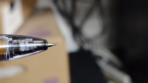 A close-up view of a ballpoint pen tip with the internal mechanism visible. The background is blurred, emphasizing the pen.