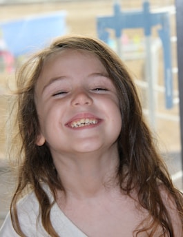 A child with long brown hair is smiling widely, showing their teeth. The background appears to be blurred, possibly indicating an outdoor setting with play equipment.